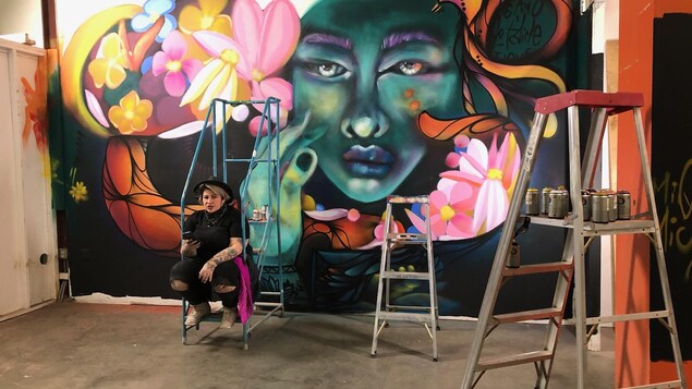 Mique Michelle est assise sur un escabot devant une murale qui dépeint une femme entourée de fleurs et de couleurs. En avant-plan : des cannettes de peinture à aérosol et d'autres escabots.
