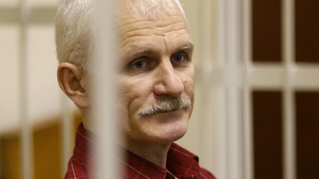 Portrait d'Ales Bialiatski derrière les barreaux d'un box d'accusés. 