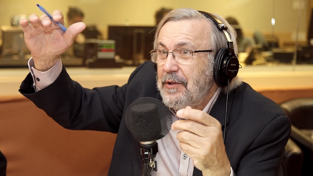 Yanick Villedieu parle dans un micro de radio en levant les bras dans les airs.