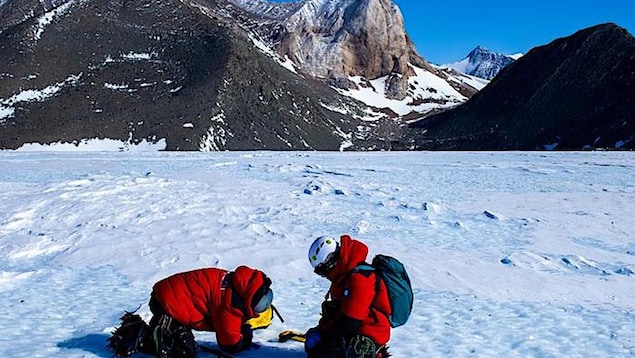 Deux chercheurs agenouillés échantillonnent la glace au pied d'une chaîne de montagnes de l'Antarctique