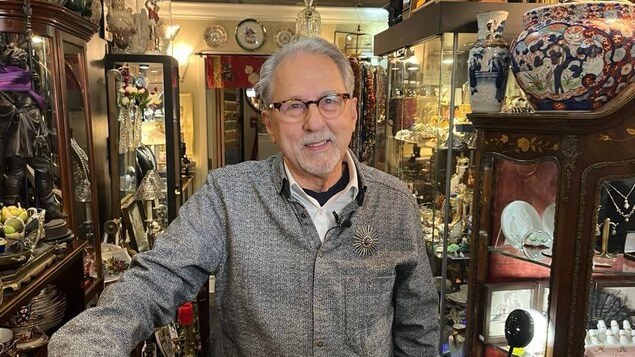 Un homme aux cheveux blanc pose dans un magasin d'antiquités rempli de meubles, de vaisselle et d'objet de collection 