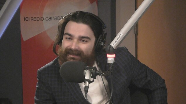 Un homme barbu avec les cheveux bruns sourit lors d'une entrevue dans un studio de Radio-Canada au Manitoba.