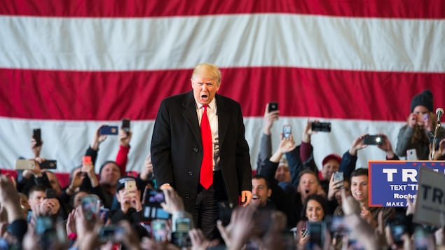 Donald Trump harangue la foule lors d'un événement électoral, devant un immense drapeau américain.
