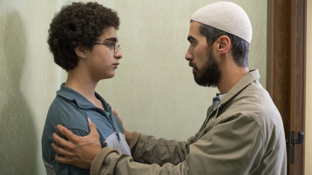 Un homme coiffé d'un kufi fait face à un jeune garçon et lui prend les épaules.
