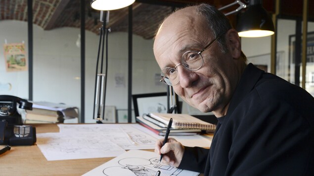 Philippe Geluck est assis à une table en train de dessiner et il se tourne vers le photographe.