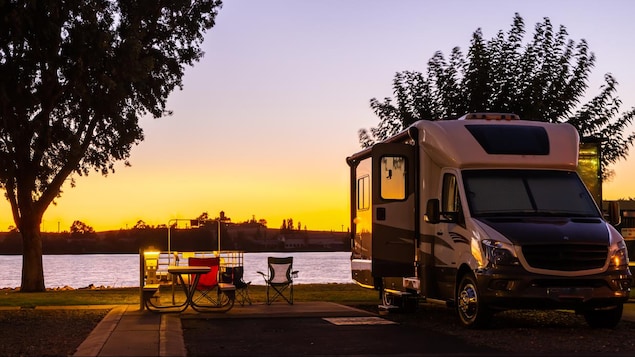 Photo prise au coucher du soleil d'une véhicule récréatif stationné au bord d'un plan d'eau avec des chaises et une table de camping sorties. 