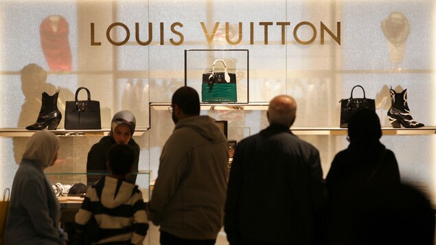 Des gens passent devant une boutique Louis Vuitton et regardent les souliers et les sacs à main présentés dans la vitrine.
