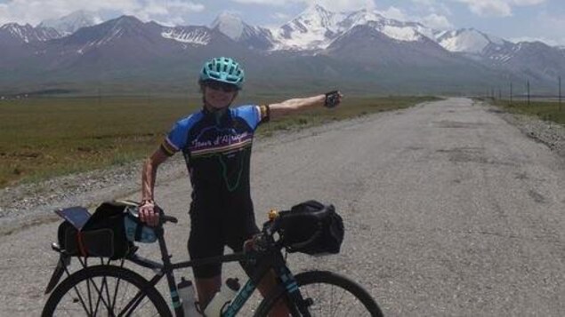 Brenda Trentholme a une main sur le sièege de son vélo et elle pointe les montagnes enneigées en arrière-plan avec son autre main.