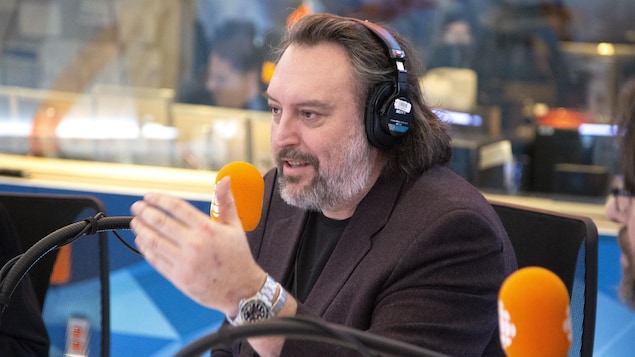 Assis derrière un micro, il lève la main gauche en parlant.