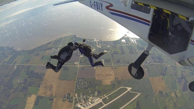 Deux parachutistes en vol libre entre le sol manitobain et l'avion qu'ils viennent de laisser.