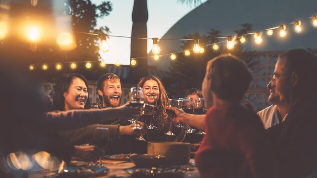 Des amis se font un toast avec des coupes de vin. Ils sont tous assis autour d'une table à l'extérieur, en début de soirée.