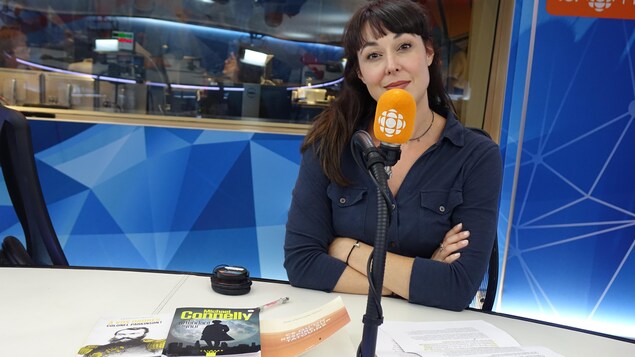 Une femme présente trois livres à la caméra, les bras croisés devant un micro orange dans un studio de radio. 