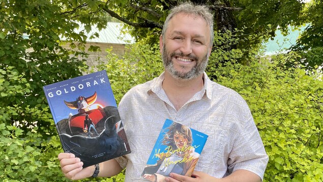 Dany Côté souriant devant des arbres et tenant deux bandes dessinées dans ses mains.