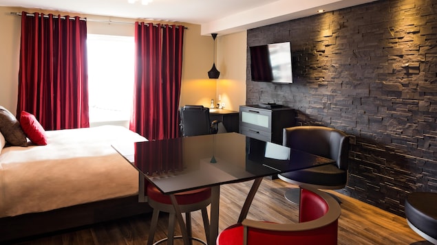 Chambre d'hôtel avec lit, rideau rouge, table, chaises et mur de pierre.