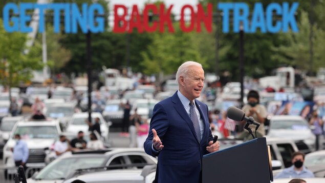 Avec en arrière-plan des lettres géantes disant « Remettre l'Amérique sur les rails », Joe Biden, derrière un lutrin, parle devant plusieurs automobilistes.