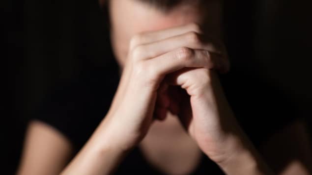 Une femme dont le visage est caché par ses mains en premier plan, paume droite sur poing gauche. 