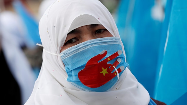 Une manifestante qui porte un masque protecteur contre la COVID-19 sur lequel on peut voir une main rouge aux couleurs du drapeau chinois proteste contre le traitement des Ouïgours par la Chine.