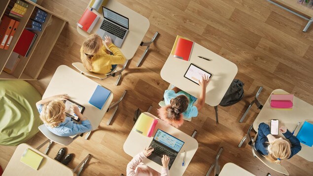 Une vue de haut d'une classe primaire où les enfants sont assis devant différents écrans; ordinateur, tablettes ou cellulaires.