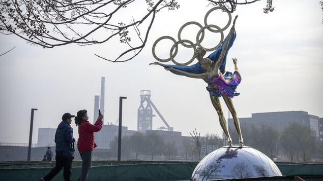 Une femme photographie une statue de patineurs artistiques surmontée des anneaux olympiques devant un paysage industriel. 