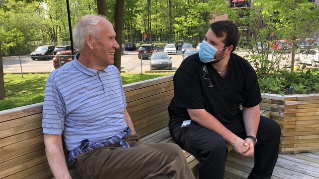 Une personne aîné assis sur un banc en compagnie d'un jeune homme portant le masque.