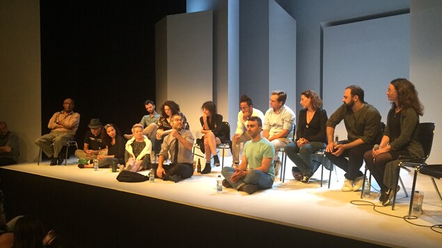 14 membres de la troupe de de la pièce de théâtre « À te regarder ils s’habitueront »assis sur une scène devant un public lors d'une discussion