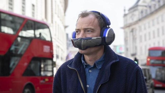 Un homme marche dans les rues de Londres avec un casque d'écoute circum-aural sur la tête : une large bande rectangulaire contenant les filtres à air couvre sa bouche. 