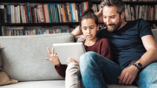Une jeune adolescente montre quelque chose à son père sur une tablette. Les deux sont assis sur un fauteuil gris.