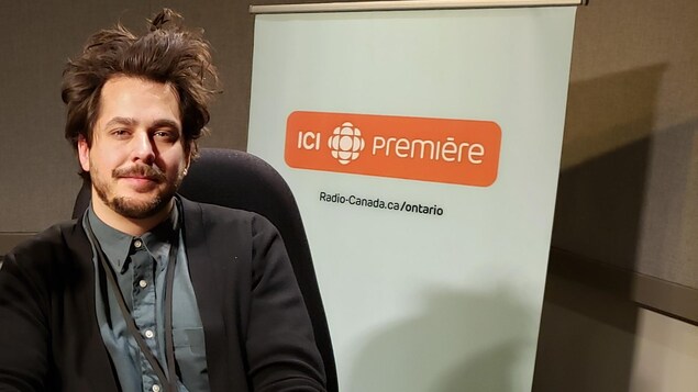 L'auteur-compositeur-interprète Matt Holubowski, assis en studio devant une bannière ICI Première