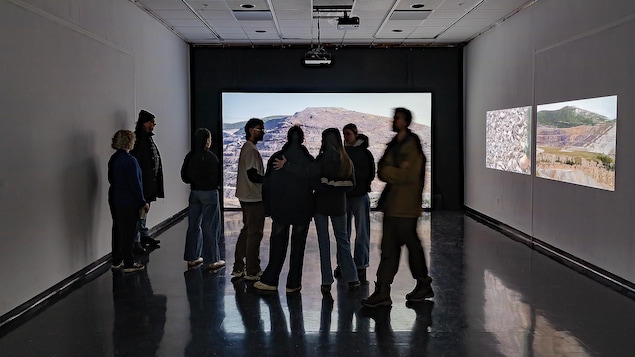 Des personnes dans une galerie où il y a trois projections sur les murs.