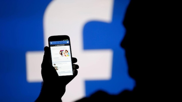 Une personne à contre-jour tient un téléphone ouvert sur Facebook. En fond, le f de Facebook sur un grand écran.