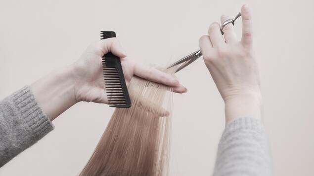 Une coiffeuse tient un peigne dans une matin, un ciseau dans l'autre et s'apprête à couper une mèche de cheveux châtains. 