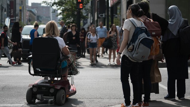 Des piétons et une dame en fauteuil roulant attendent à une intersection de la rue Sainte-Catherine.