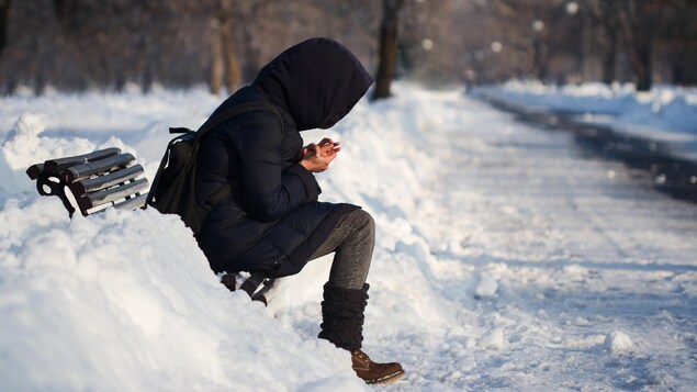Une femme, le capuchon de son manteau sur la tête, est assise seule sur un banc de parc une journée d'hiver. 