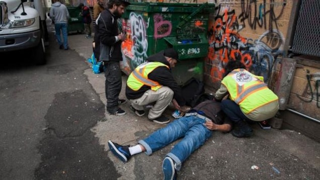 Des secouristes sont accroupis pour traiter un homme étendu au sol, sur le dos, dans une ruelle dont les murs sont couverts de graffiti.