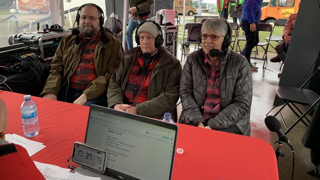 Deux hommes et une femme vêtus d'une chemise à carreaux sont en entrevue radio