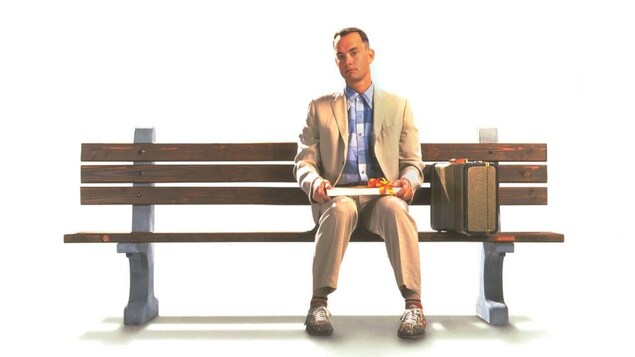 Un homme est assis sur un banc public avec une boite de chocolat sur les genoux. 