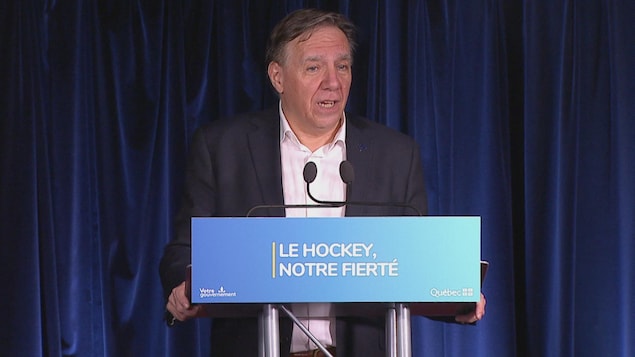 Le premier ministre du Québec, en veston, parle pendant une conférence de presse, devant un podium où il est inscrit « Le hockey, notre fierté ». 