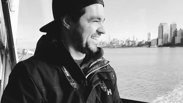 Souriant, Zacharry-David Dufour se trouve sur un bateau et tient un appareil photo dans ses mains.