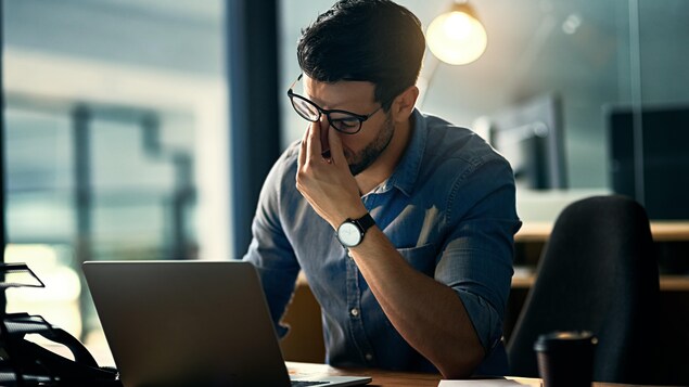 Un an après le début de la pandémie, l'entreprise Morneau Shepell a publié un rapport qui montre que le sentiment d'isolement n'a jamais été aussi fort depuis le début de la pandémie. Un homme se tient le nez en signe d'épuisement devant son ordinateur portable posé sur une table.