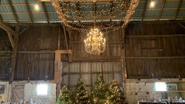 Un immense chandelier est suspendu du plafond d'une grange qui est toute décorée avec des sapins de Noël illuminés