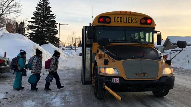 Dans un quartier résidentiels, en hiver, trois enfants montent à bord d’un autobus scolaire.
