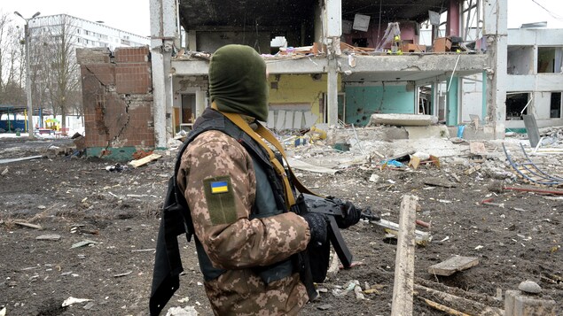 Un soldat cagoulé et armé regarde un immeuble éventré.