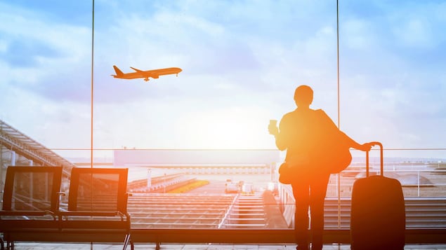 Une femme se tient devant la baie vitrée d'un aéroport pendant qu'un avion décolle sur la piste devant elle.