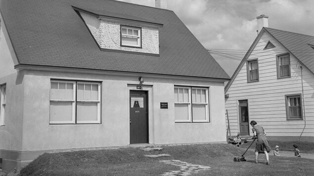Une femme tond une pelouse devant une maison alors que trois enfants sont assis.