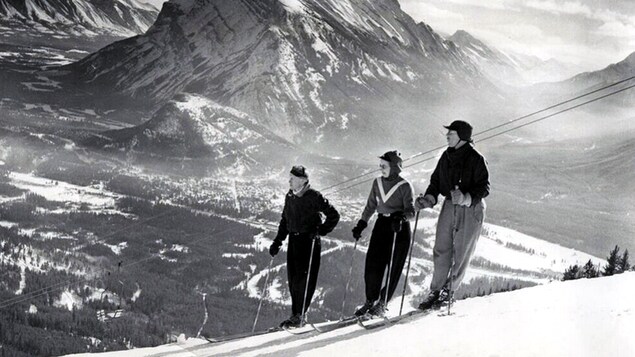 Trois skieurs sur une pente de ski avec une montagne majestueuse en arrière-plan. 