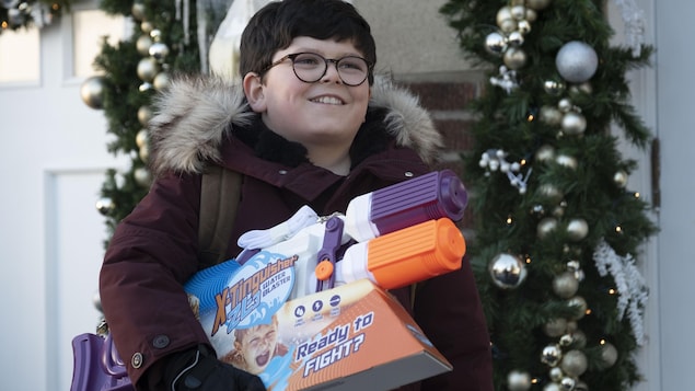 Un garçon de 10 ans, vêtu d'un manteau d'hiver et tenant un pistolet à eau sous le bras, sourit depuis l'extérieur d'une maison, décorée pour le temps des Fêtes. 