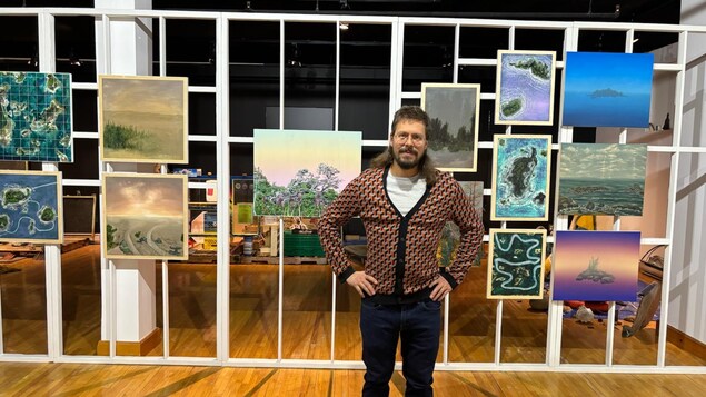 Un homme debout devant des tableaux qu'il a lui-même peints.