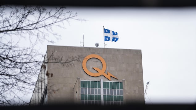 Gros plan sur l'enseigne lumineuse du logo d'Hydro-Québec installée au sommet du bâtiment du siège social de la société d'État près d'un drapeau du Québec qui flotte au vent.