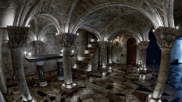 Un sous-sol avec un plancher en pierres sombres, des colonnes et des lumières. Le tout dans une ambiance plutôt lugubre.