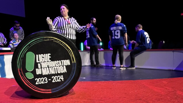 Au premier plan un cercle avec l'inscription «Ligue d'Improvisation du Manitoba 2023-2024». Au second plan, des joueurs de la LIM et des arbitres.